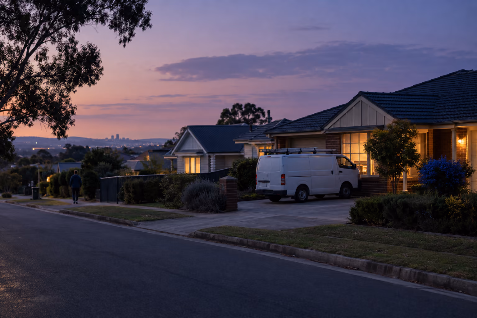 A suburban Australian street at dusk, with lights coming on in the houses and a plumber's van parked in a driveway
