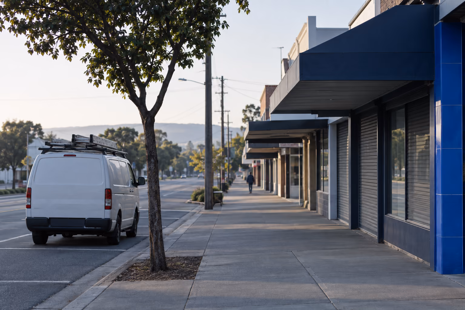 An electrician's van parked outside a row of suburban shopfronts in the early morning, awnings and footpath empty