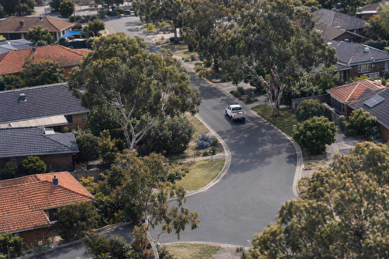 An aerial view of an Australian suburb of red and grey rooftops and gum trees, with a tradesperson's ute turning into a quiet street