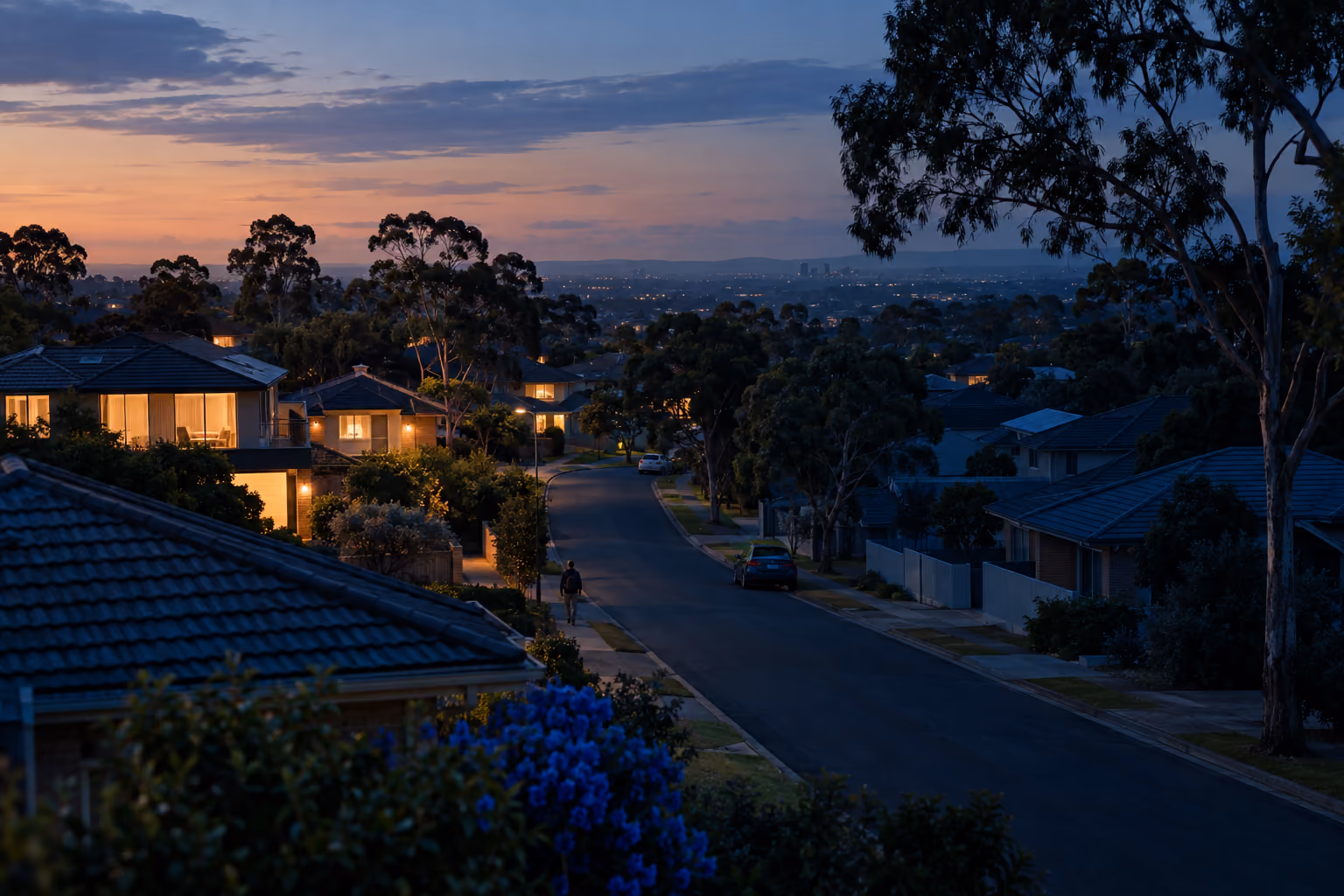 An Australian suburban street at dusk seen from slightly above, with half the houses lit and half in shadow