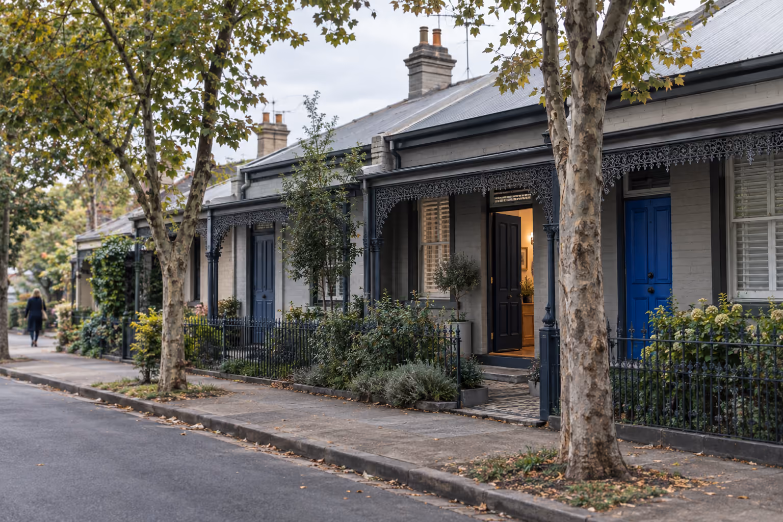 A row of terrace houses in an inner Australian suburb, each front door a different shade of blue, one door open with warm light inside