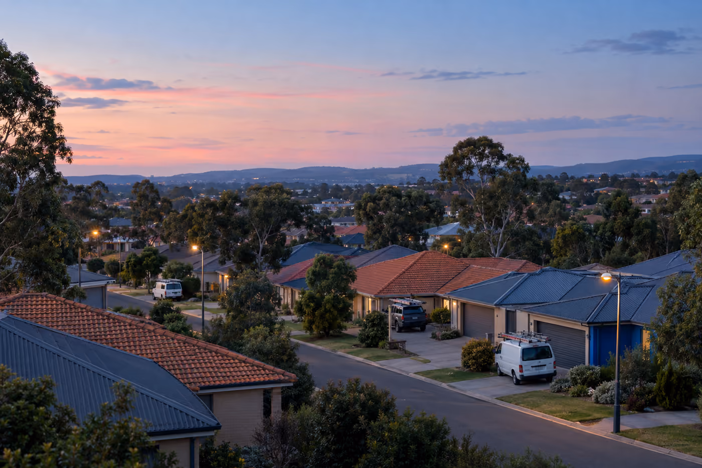 Rows of suburban Australian rooftops in terracotta and steel at dusk, seen from a low rise, with a few work vans in driveways